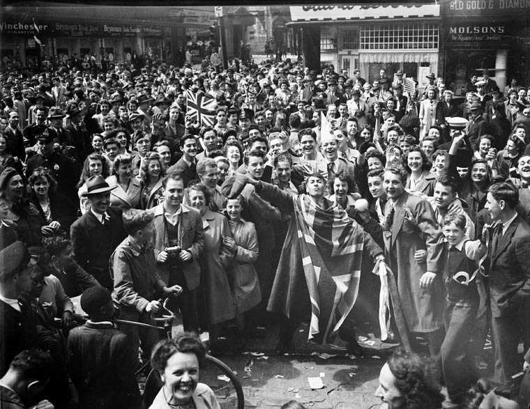 Black and white photograph. With storefronts in the background at an intersection, the streets are packed with people, several with bicycles, most facing toward the camera. Their faces are jubilant, and many wave flags. Hands are raised in celebration.